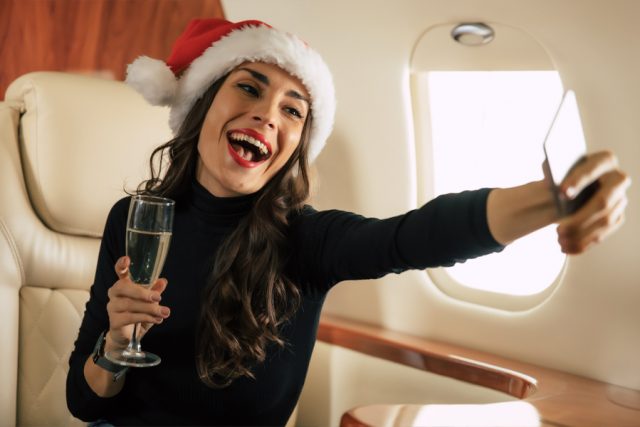 Lady on jet with Santa hat on, holding a glass and taking a selfie.